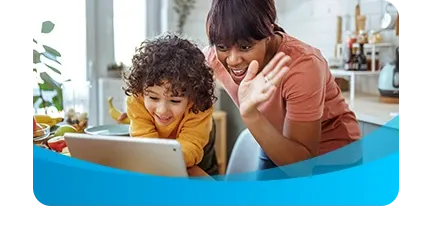 A smiling mother waves at a tablet screen while a young child watches during a video call in the kitchen.